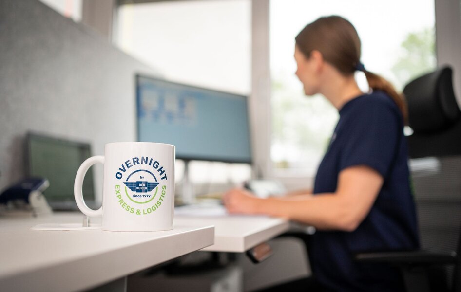 Overnight Express & Logistics – Kundenservice im Büro Tasse mit Overnight Express & Logistics Logo auf einem Schreibtisch, im Hintergrund eine Mitarbeiterin am Büroarbeitsplatz