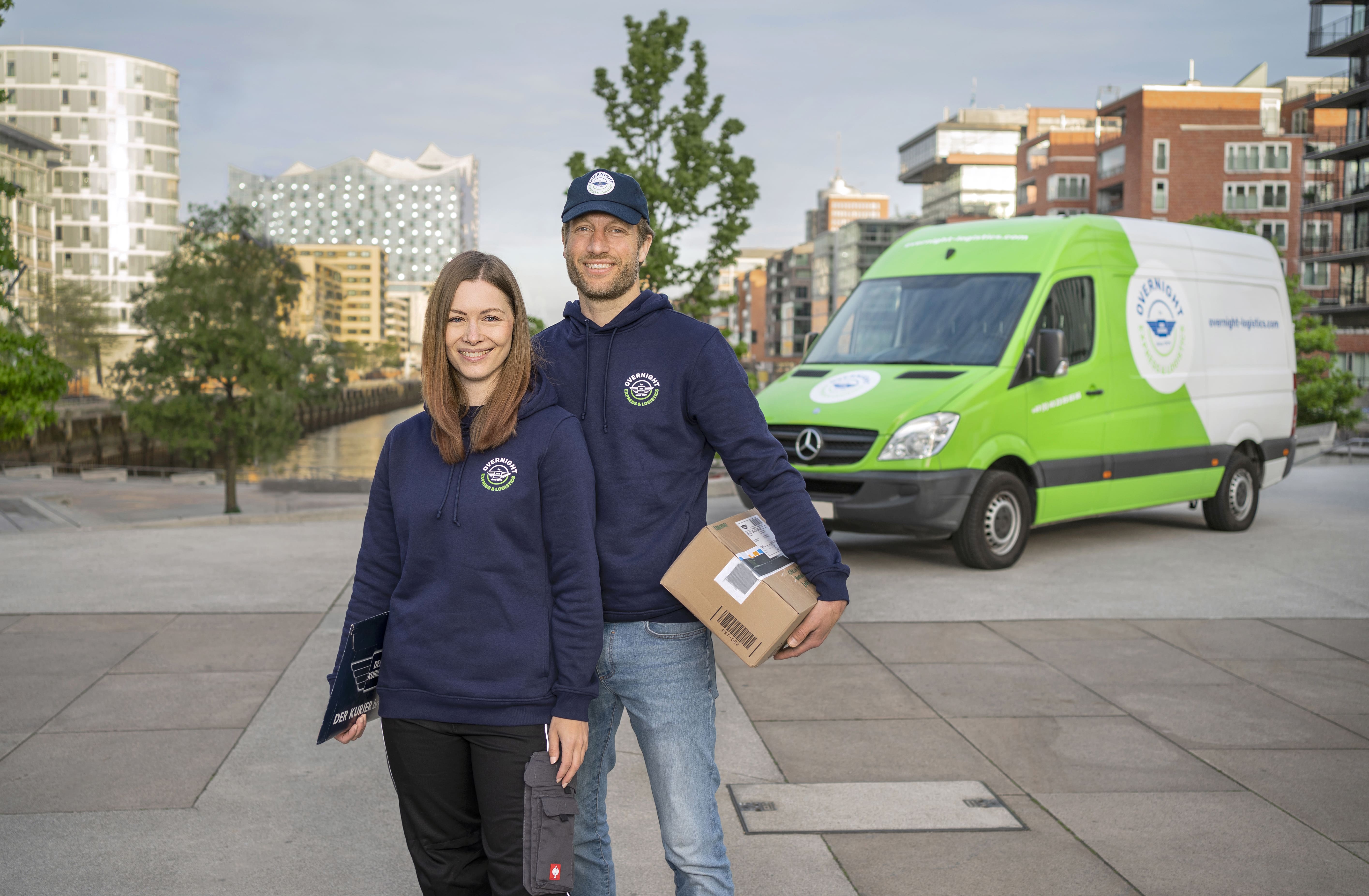 Ein Mann und eine Frau stehen lächelnd vor einem Lieferwagen, beide in blauen Kapuzenpullis. Der Mann hält ein Paket in der Hand.